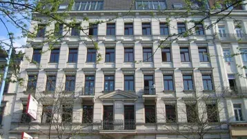 Klassisches Mehrfamilienhaus mit heller Fassade, vielen Fenstern und französischen Balkonen, umgeben von frühlingshaften Bäumen.