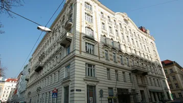 Ornates Mehrfamilienhaus an einer Straßenecke mit vielen Fenstern und Balkonen unter blauem Himmel.
