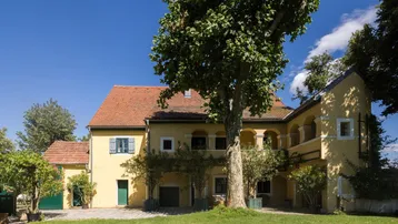 Historisches Gebäude mit rotem Ziegeldach, umgeben von einem gepflegten Rasen und hohen Bäumen.