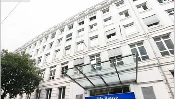 Weißes Bürogebäude mit vielen Fenstern, Jalousien und einem gläsernen Vordach über dem Eingang.
