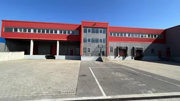 Modernes Industriegebäude mit roter Fassade, Laderampen und großem gepflasterten Vorplatz unter blauem Himmel.
