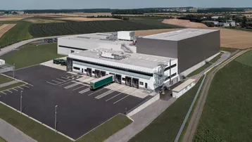 Luftaufnahme eines großen Industriegebäudes mit Laderampen, Parkplätzen und umgebenden Feldern.