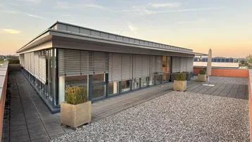 Große Dachterrasse mit Kiesfläche, Fliesenwegen und modernen Pflanzkübeln, umgeben von einem Gebäude mit Jalousien.