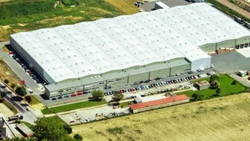 Luftaufnahme eines großen Industriegebäudes mit weitläufigen Parkplätzen und umliegenden Feldern.