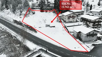 Luftaufnahme eines schneebedeckten Grundstücks mit roter Umrandung, umgeben von Wohnhäusern und Wald, mit einer Straße und einem Fluss im Vordergrund.