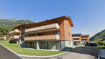 Modernes Mehrfamilienhaus in Holz- und Steinfassade mit Balkonen und Bergblick, umgeben von grüner Landschaft.