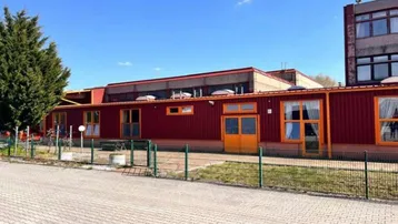 Gebäude mit roter Fassade, großen Fenstern und einem grünen Zaun im Vordergrund unter blauem Himmel.