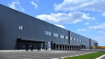 Moderne Logistikhalle mit zahlreichen Laderampen und großem Parkplatz unter blauem Himmel.