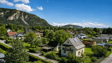 Luftaufnahme einer Wohnsiedlung mit Einfamilienhäusern, umgeben von grünen Wäldern und Bergen.