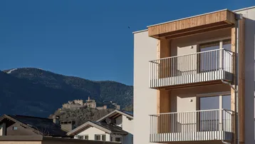 Moderne Wohnanlage mit Holzverkleidung und Balkonen, im Hintergrund eine Burg vor Bergkulisse.