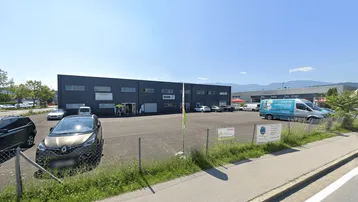Gebäudeansicht eines modernen Gewerbeobjekts mit Parkplatz und Bergen im Hintergrund unter blauem Himmel.