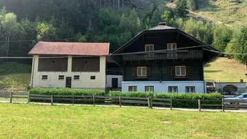 Traditionelle Gebäude in ländlicher Umgebung mit bewaldeten Hügeln im Hintergrund und grüner Wiese im Vordergrund.