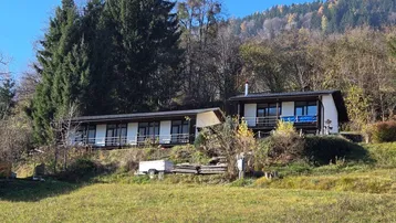 Zwei Gebäude in einer grünen Hügellandschaft mit Wald im Hintergrund unter blauem Himmel.