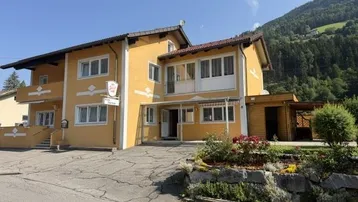 Gelbes Gebäude mit traditioneller Architektur, umgeben von Bergen und grüner Landschaft unter blauem Himmel.