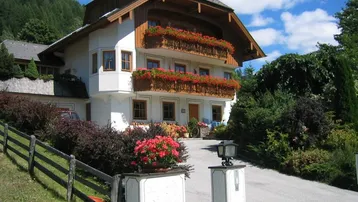 Traditionelles Haus im alpinen Stil mit blühenden Blumenkästen an den Holzbalkonen und grüner Umgebung.