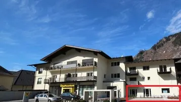 Mehrstöckiges Gebäude mit Balkonen und Geschäftsräumen im Erdgeschoss, umgeben von Bergen unter blauem Himmel.