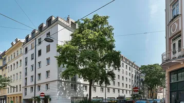 Mehrstöckiges Wohngebäude mit weißer Fassade und grünen Bäumen an einer städtischen Straße.