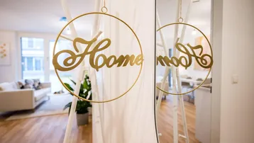 Goldene "Home"-Dekoration im Vordergrund, mit hellem Wohnzimmer im Hintergrund.