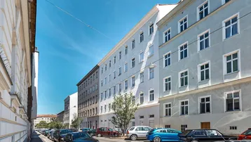 Weitläufige Straßenansicht mit mehreren Wohngebäuden und parkenden Autos unter blauem Himmel.
