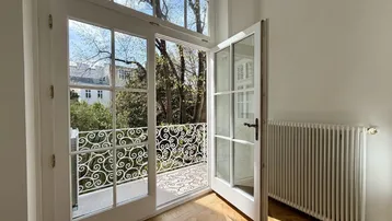 Heller Raum mit Fischgrätparkett und Zugang zu einem Balkon mit schmiedeeisernem Geländer und Blick ins Grüne.