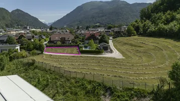 Luftaufnahme eines Baugrundstücks mit umliegenden Häusern, Feldern und Bergpanorama.