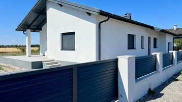 Modernes Einfamilienhaus mit weißer Fassade, dunklem Zaun und Rollläden unter blauem Himmel.