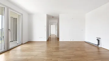 Helles, leeres Wohnzimmer mit Holzboden und Zugang zu einem Balkon oder einer Loggia.