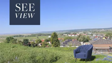 Weitläufiger Blick über grüne Felder und ein Dorf mit einem See in der Ferne unter blauem Himmel.