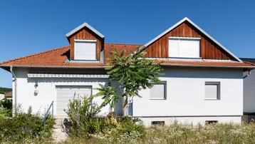 Einfamilienhaus mit weißer Fassade, rotem Ziegeldach und Dachgauben in ländlicher Umgebung.