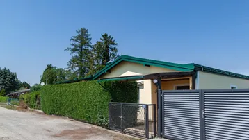 Einfamilienhaus mit gelber Fassade, grüner Hecke und modernem Eingangstor an einem sonnigen Tag.