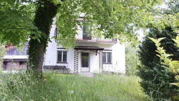 Weißes Haus mit Balkon und üppiger Vegetation im Vordergrund, umgeben von Natur.