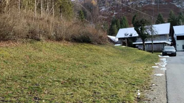 Blick auf eine grüne Wiese mit leichtem Schneefall, umgeben von bewaldeten Hügeln und ländlichen Gebäuden.