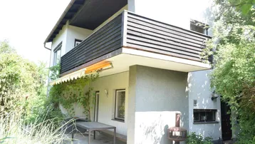 Hausfassade mit Balkon und einer möblierten Terrasse unter einer Markise, umgeben von Grün.