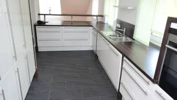 Moderne Küche mit weißen Hochglanzfronten, dunkler Arbeitsplatte und anthrazitfarbenen Fliesenboden.