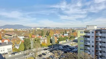 Weitläufiger Blick über die Stadt mit Wohngebäuden, Parkplätzen und grünen Hügeln.