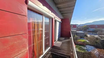 Sonniger Balkon mit Holzboden, roter Fassade und weitem Blick auf die Umgebung.