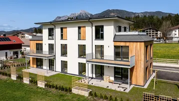 Moderne Wohnanlage mit weißer Fassade, Holzverkleidung und Balkonen vor alpiner Bergkulisse.