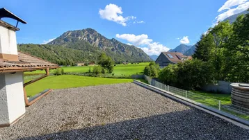 Weitläufiger Blick von einer Terrasse über grüne Felder und majestätische Berge unter blauem Himmel.
