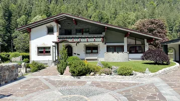 Traditionelles Alpenhaus mit gepflasterter Auffahrt, gepflegtem Vorgarten und bewaldetem Berghintergrund.