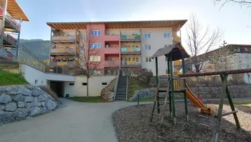 Mehrfamilienhaus mit bunten Balkonen, Spielplatz im Vordergrund und Bergblick.