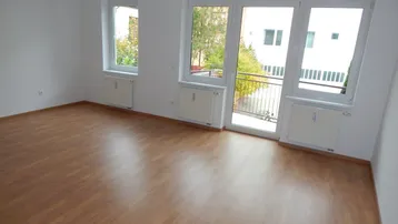 Helles, leeres Zimmer mit Laminatboden und Zugang zu einem kleinen Balkon.