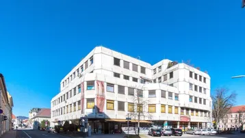 Mehrstöckiges Geschäftsgebäude mit weißer Fassade und vielen Fenstern an einer belebten Straße unter blauem Himmel.