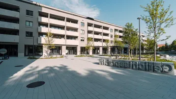 Modernes Mehrfamilienhaus mit Balkonen und einer gepflasterten Außenfläche, umgeben von jungen Bäumen.