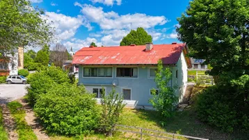 Zweistöckiges Wohnhaus mit rotem Ziegeldach und umgebender Grünfläche unter blauem Himmel.