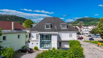 Gebäudeansicht mit moderner Glasfassade und umliegender Landschaft unter blauem Himmel.