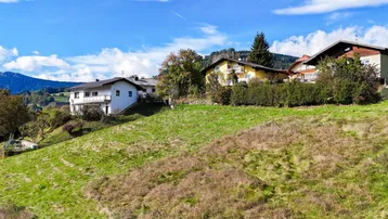Mehrere Wohnhäuser auf einem sonnigen Hügel mit grüner Wiese und bewaldeten Bergen im Hintergrund.