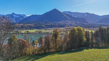 Panoramablick auf einen See mit umliegenden Bergen und herbstlicher Vegetation unter blauem Himmel.