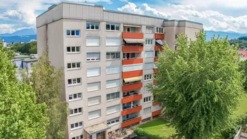 Luftaufnahme eines Mehrfamilienhauses mit Balkonen, grüner Umgebung und Bergblick im Hintergrund.