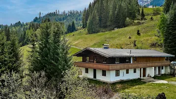 Traditionelles Alpenhaus mit Holzbalkon, umgeben von grünen Hügeln und Nadelbäumen, mit Bergblick.