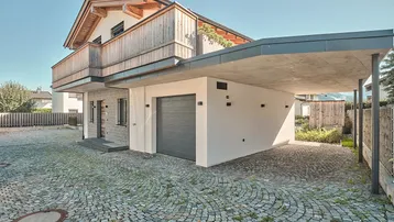 Modernes Haus mit Garage, Carport und gepflastertem Vorplatz unter blauem Himmel.